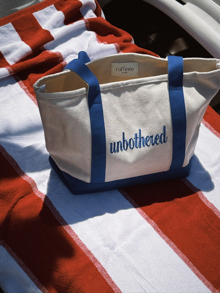 White tote bag with blue handles and 'unbothered' text on a red and white striped surface.